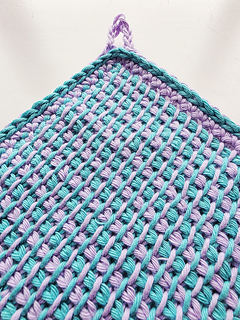 Close-up of Tunisian crocheted square cloth with hanging loop, worked in a turquoise and lavender pattern on a white background.