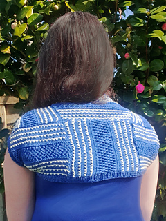 Woman wearing a blue and white knitted shrug stands facing away in a sunny garden, with a leafy background.