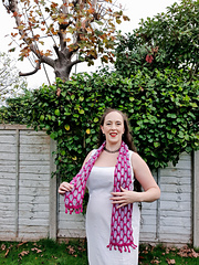 Smiling woman in a white dress with a pink and light blue crochet scarf stands in a garden with green hedges and a tree. She wears a black necklace.