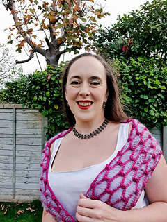 Smiling woman in a white dress with a pink and light blue crochet scarf stands in a garden with green hedges and a tree. She wears a black necklace.