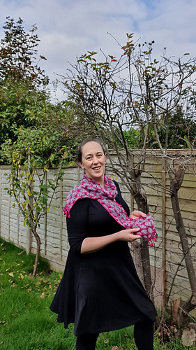 Smiling woman in a black dress with a pink and light blue crochet scarf stands in a garden with green hedges and a tree.