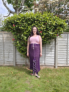 A woman in a pink top and long purple crocheted ruffle edged split skirt stands on grass, backed by a garden fence and lush green hedge with flowers. Overcast sky above.