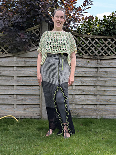 A woman in a green crocheted top and long grey, yellow and black crocheted ruffle edged split skirt stands on grass, backed by a garden fence 