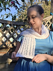 Woman in a blue dress smiles while holding up a white lace knitted scarf with pink ribbons woven through in a leafy garden.
