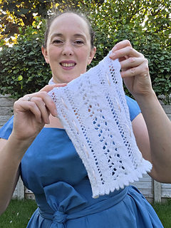 Woman in a blue dress smiles while holding up a white lace knitted scarf with pink ribbons woven through in a leafy garden.