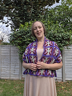 crochet lace shawl with colourful butterfly design  in blues purples, pinks and green on a woman in a leafy garden (close, front)