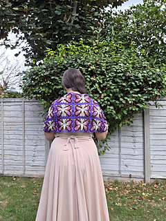 crochet lace shawl with colourful butterfly design  in blues purples, pinks and green on a woman in a leafy garden (back)