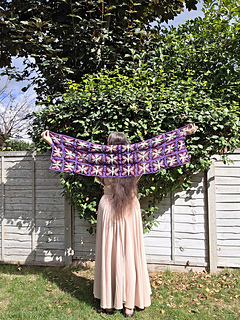 crochet lace shawl with colourful butterfly design  in blues purples, pinks and green held up behind a woman in a leafy garden (back open)