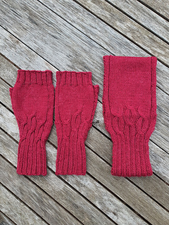 Pair of deep red cable knitted fingerless gloves shown with matching headband (not included in Mitts pattern) lying on a weathered grey wooden slatted table.