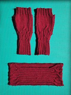 Pair of deep red cable knitted fingerless gloves shown with matching headband (not included in Mitts pattern) pinned on a green foam mat.