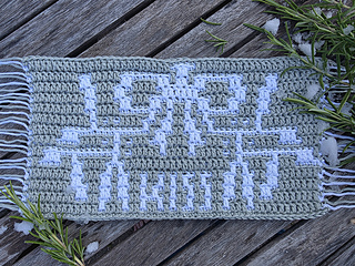 Crocheted green and white mosaic panel with spray of mistletoe tied with a bow and text spelling KISS in white on a weathered wooden deck, with rosemary sprigs and melting snow.