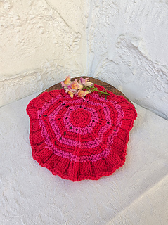 Red knitted circular washcloth with pink stripes with flowers on a wooden stand against a white textured wall.