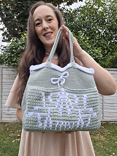Smiling woman outdoors holds a pale sage green crocheted tote with mistletoe design and KISS text in white, standing before a leafy fence and trees.