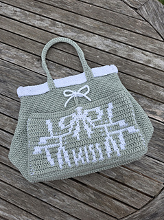 A pale sage green crocheted tote with mistletoe design and KISS text in white, on a wooden slatted table