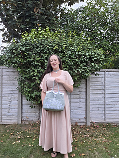 Smiling woman outdoors holds a pale sage green crocheted tote with mistletoe design and KISS text in white, standing before a leafy fence and trees.
