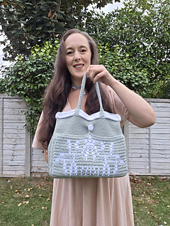 Smiling woman outdoors holds a pale sage green crocheted tote with mistletoe design and KISS text in white, standing before a leafy fence and trees.