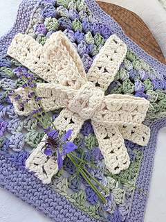 Crocheted lavender, green, and cream granny square on a wooden board, decorated with purple wildflowers and a cream crocheted ribbon