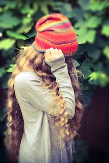 Woman with long blonde hair wearing a bright red slouchy knit hat with the ribbed brim folded up and her hand in front of her face.