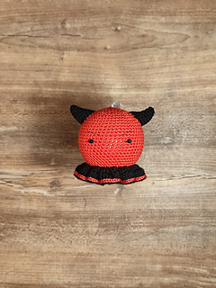 Small crocheted devil Christmas bauble with red body, tiny horns, pointed tail, and a mischievous smile. Put on the table.