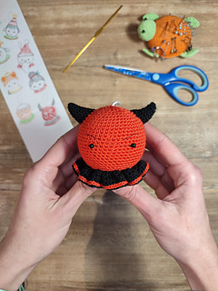 Small crocheted devil Christmas bauble with red body, tiny horns, pointed tail, and a mischievous smile. Held in the hands, crochet tools at the table in the background.