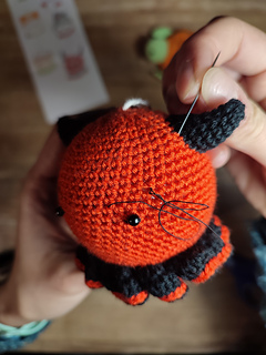 Small crocheted devil Christmas bauble with red body, tiny horns, pointed tail, and a mischievous smile. Embroidering the eyebrows above the eyes.