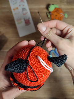Small crocheted devil Christmas bauble with red body, tiny horns, pointed tail, and a mischievous smile. Sewing the horns onto the head.