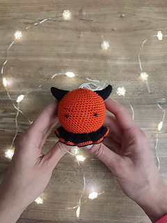 Small crocheted devil Christmas bauble with red body, tiny horns, pointed tail, and a mischievous smile. Held in the hands, Christmas lights at the table in the background.