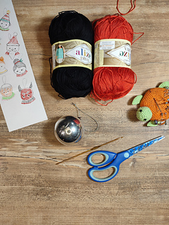 Small crocheted devil Christmas bauble with red body, tiny horns, pointed tail, and a mischievous smile. Crochet tools at the table in the background.