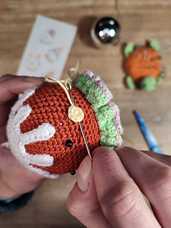 Crocheted gingerbread brown head, white icing details, and colorful decorative buttons. Sewing on the cheeks, crochet tools put on the table, scissors, crochet hook, plain Christmas bauble.