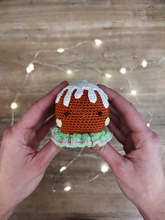 Crocheted gingerbread brown head held in the hands, white icing details, and colorful decorative buttons. Christmas lights in the background on the table.