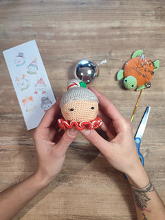 Crochet Mrs. Claus doll wearing a red collar with white details and gray hair in a bun. Held in the hands, crochet tools on the table in the background, scissors, crochet hook, pins.