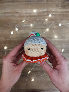 Crochet Mrs. Claus doll wearing a red collar with white details and gray hair in a bun. Held in the hands, Christmas lights in the background on the table.