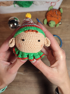 Handmade crochet Christmas elf with pointed hat, striped outfit, and playful expression. Held in the hands. Crochet tools visible in the background.