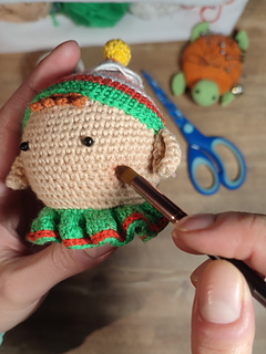 Handmade crochet Christmas elf with pointed hat, striped outfit, and playful expression. Held in the hands. Adding a face blush onto the cheeks.