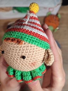 Handmade crochet Christmas elf with pointed hat, striped outfit, and playful expression. Held in the hands. Crochet tools visible in the background. Detailed face and head view.