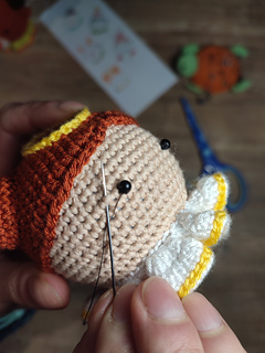 Crochet amigurumi style Christmas bauble. Brown hair with two buns, yellow halo at the top of the head and white-yellow collar. Embroidering the eyelashes onto the angel's face.