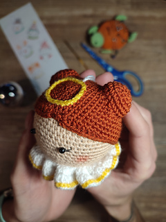 Crochet amigurumi style Christmas bauble. Brown hair with two buns, yellow halo at the top of the head and white-yellow collar. Held in the hand, left side face detail. Crafting tools are on the table in the background, Christmas bauble, scissors, hook, pins and hand drawn sketches.