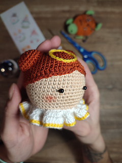Crochet amigurumi style Christmas bauble. Brown hair with two buns, yellow halo at the top of the head and white-yellow collar. Held in the hand, right side face detail. Crafting tools are on the table in the background, Christmas bauble, scissors, hook, pins and hand drawn sketches.