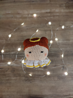 Crochet amigurumi style Christmas bauble. Brown hair with two buns, yellow halo at the top of the head and white-yellow collar. Held in the hand. Christmas lights in the background on the table.