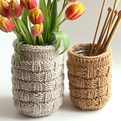 Two knitted jar cozies, one with flowers and the other with wooden knitting needles, on a light background.