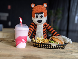 Large crochet tiger plushie sitting at a wooden table with a milkshake, burger and fries in front.