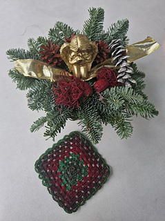 Crocheted red and green granny square coaster next to a Christmas table arrangement with fir branches, pine cones and gold decoration